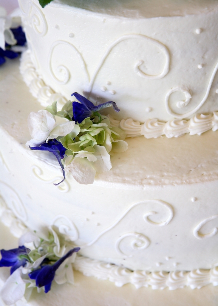 detail of a layered wedding cake with white icing and swirls and purple and white flower accents
